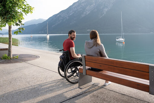  Ein Rollstuhlfahrer und eine Frau sitzen an der Seepromenade in Pertisau im Sommer und schauen auf den See.